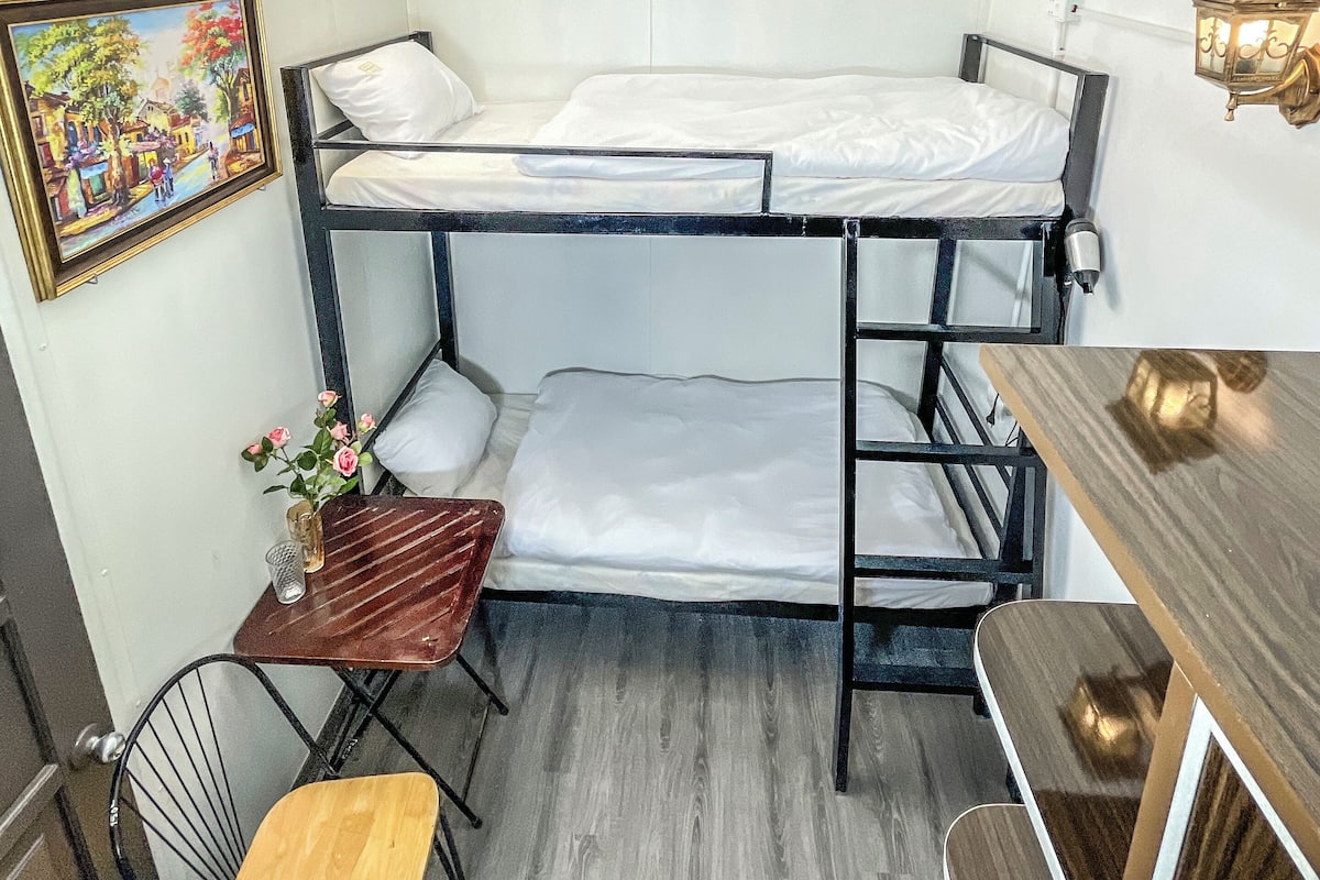 A bunk bed room with a desk and chair, a small table with a vase of flowers, and a wall-mounted lamp.