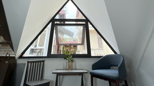 A room with a sloped ceiling, a table with chairs, a window with a view of buildings, and a vase with flowers. - HC Luxury Apartment (Hanoi)