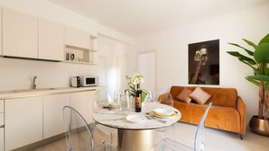 Une cuisine moderne avec des meubles blancs, une table à manger entourée de chaises transparentes, un canapé et une plante en pot.