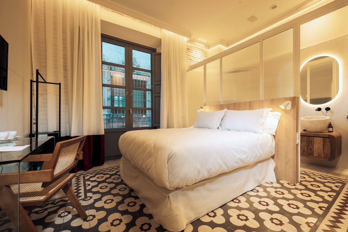 A hotel room with a large bed, a desk with a chair, a window with curtains, and a patterned floor.