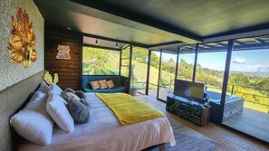 A modern bedroom with a bed, a sofa, a TV, and a balcony with a view of the countryside. - Cumbres Luxury Glamping (Guarne)