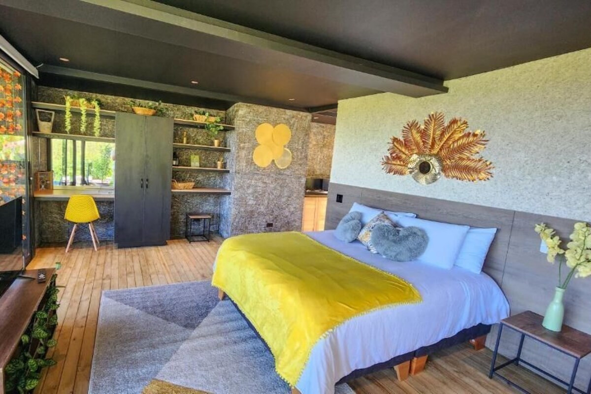 A modern bedroom with a large bed, a desk, and a shelving unit with plants.
