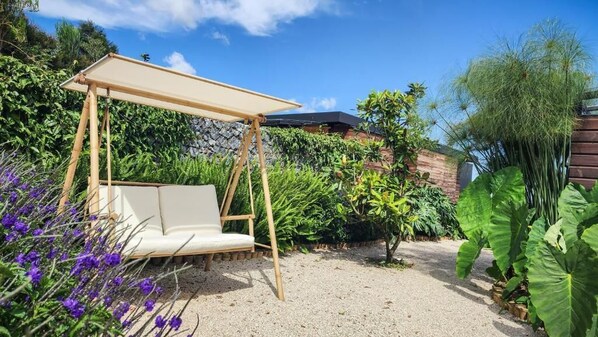 Un jardín con un columpio de madera, un banco blanco acolchado y diversas plantas.