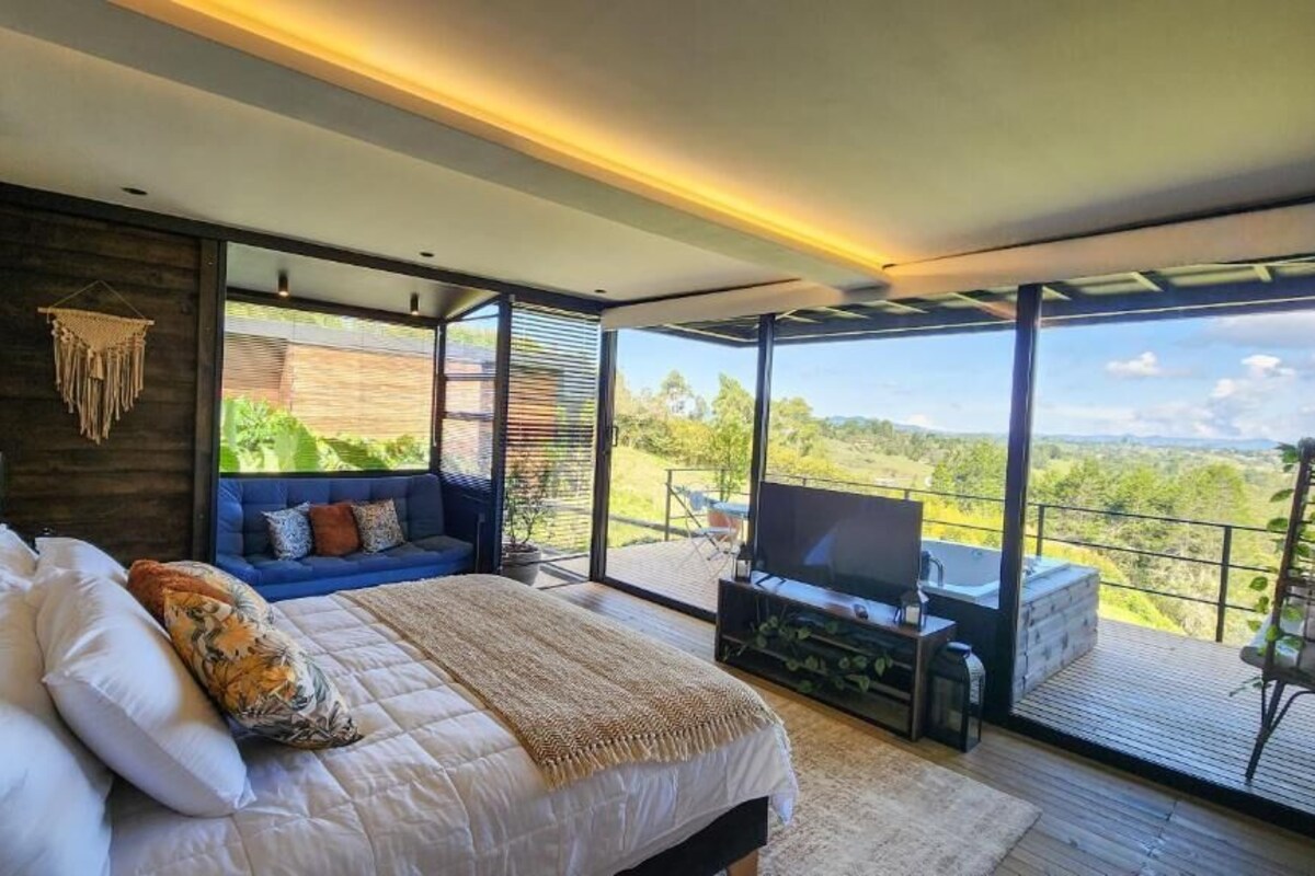 A modern bedroom with a large bed, a sofa, a TV, and a balcony with a view of the landscape.