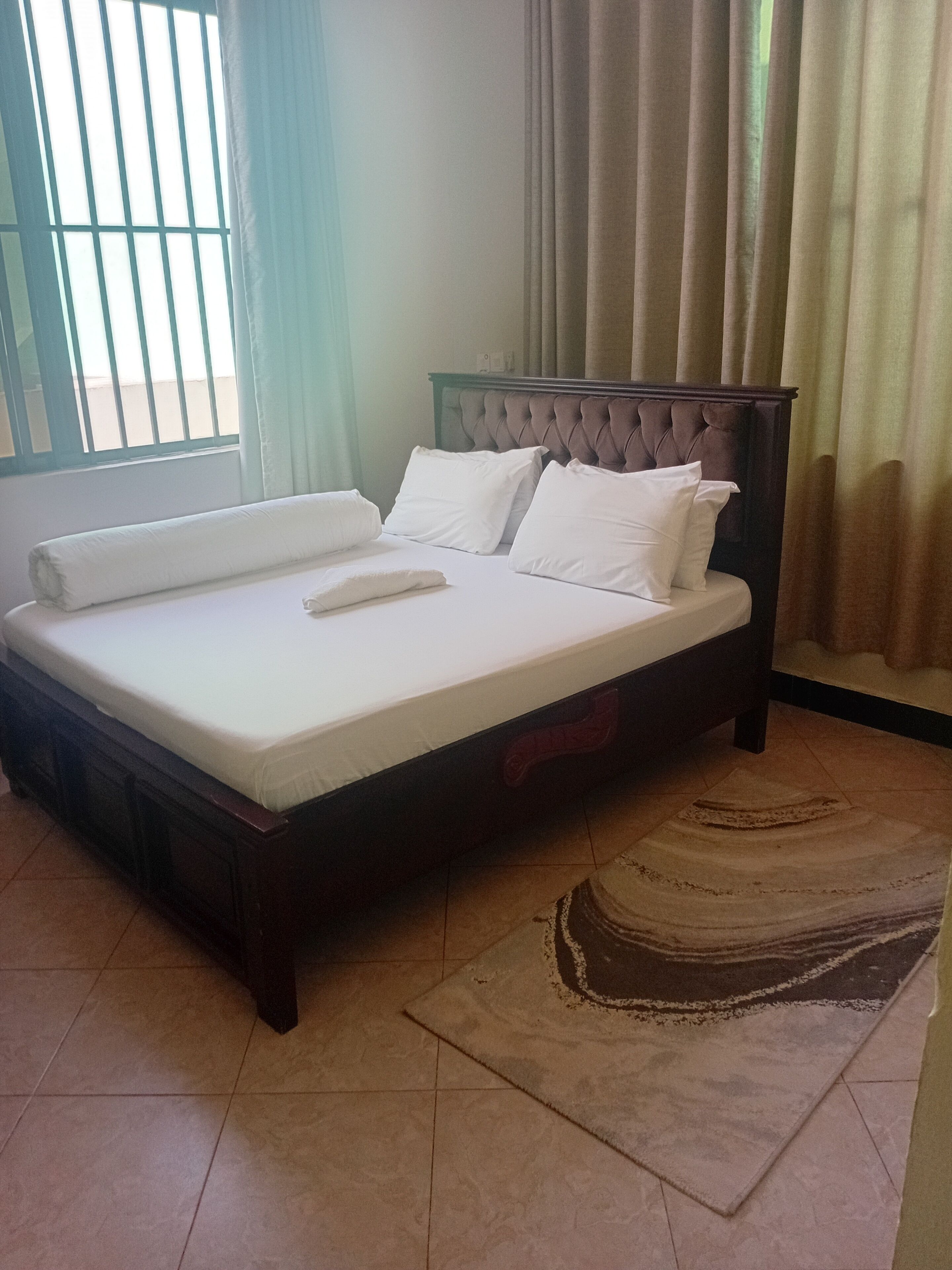 Una cama bien hecha con sábanas y almohadas blancas, un cabecero de madera oscura, y una ventana con barrotes y cortinas.