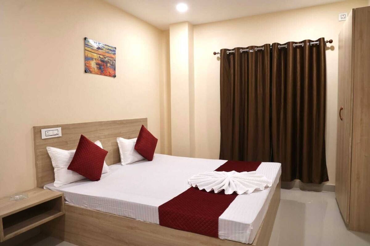 A neatly made bed with a red and white bedspread, a wooden headboard, and a red pillow. There is a nightstand with a lamp, a painting on the wall, and a brown curtain.
