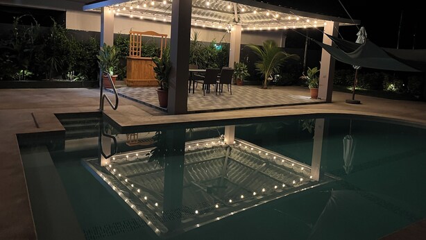 Un área junto a la piscina con una zona de estar cubierta, plantas en macetas y una hamaca.