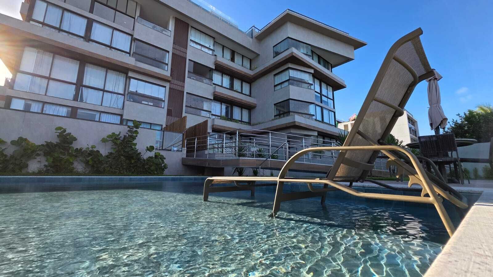 En swimmingpool med en solstol, klart vatten och en modern byggnad med balkonger i bakgrunden.