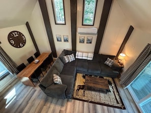 A modern living room with a sectional sofa, a wooden coffee table, and a dining area with a wooden table and chairs. There is a large wall clock and framed artwork above the sofa.