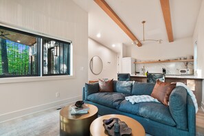 Una sala de estar moderna con un ventanal grande, un sofá azul y una cocina al fondo.