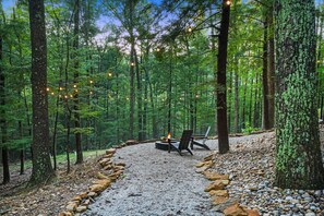 A forest campsite with a fire pit and chairs, surrounded by trees and a gravel path.