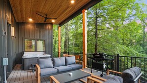 Terraza de madera con sofá, mesa de centro y zona de barbacoa, con vista a un bosque.