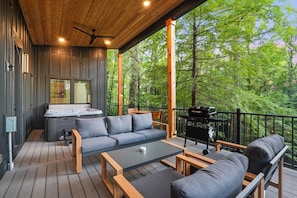 Terraza de madera con sofá, mesa de centro y zona de barbacoa, con vista a un bosque.
