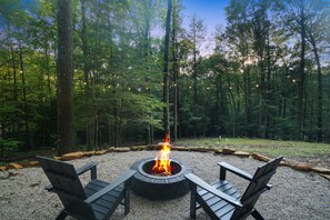 Una fogata circular con dos sillas Adirondack en un entorno boscoso.