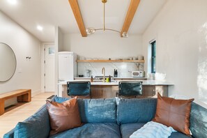 Una sala de estar moderna con un sofá azul, vigas de madera, una zona de cocina con refrigerador blanco y una mesa de comedor con sillas.