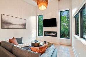 A modern living room with a large sofa, a fireplace, a flat-screen TV, and large windows offering a view of trees.