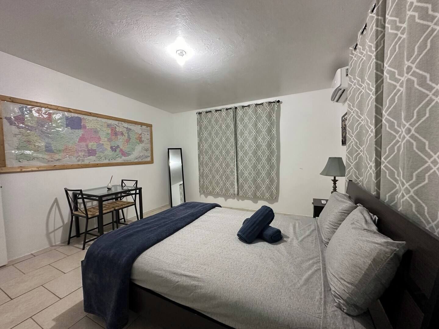 A bedroom with a bed, a desk with chairs, a map on the wall, and patterned curtains.