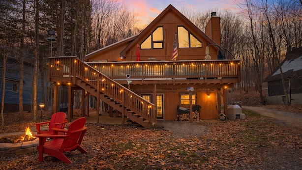 A wooden cabin with a balcony, a fire pit, and red Adirondack chairs.