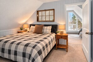 Ein Schlafzimmer mit einem großen Bett, zwei Nachttischen mit Lampen, einem Fenster mit Blick auf Bäume und einer Tür zu einem anderen Raum.