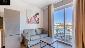 Un salon moderne avec un canapé gris, une petite table et une grande fenêtre donnant sur un port de plaisance.