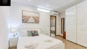 Un dormitorio moderno con una cama grande, una mesita de noche, una lámpara de noche, un cuadro enmarcado en la pared y vistas al baño.