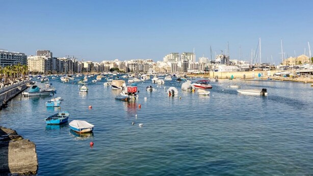 A marina with numerous boats docked, surrounded by buildings and a clear sky.
