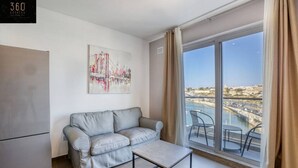 A modern living room with a grey sofa, a small table, and a large window with a view of a cityscape and water.