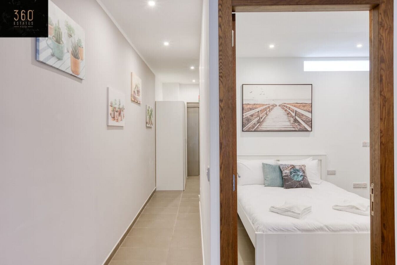A hotel room with a bed, a wooden door, and a hallway with artwork.