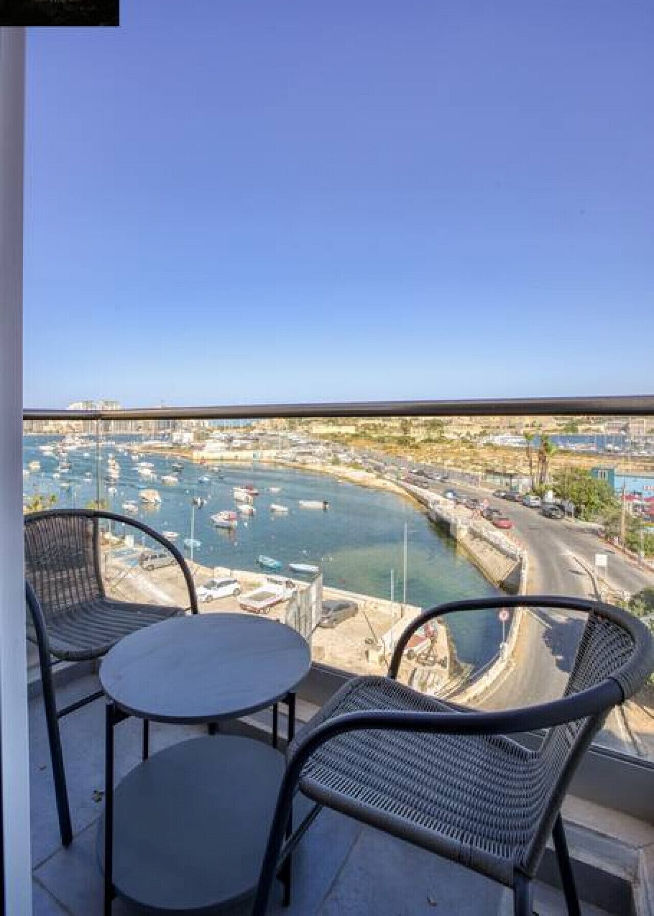 A balcony with two round tables and two wicker chairs, overlooking a marina with boats and a cityscape.