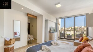A modern hotel room with a large bed, a balcony with a city view, a desk with a chair, and a small table with a lamp.