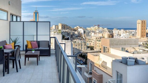 A balcony with a sofa, chairs, and a small table, overlooking a cityscape with buildings and a distant view of the sea.