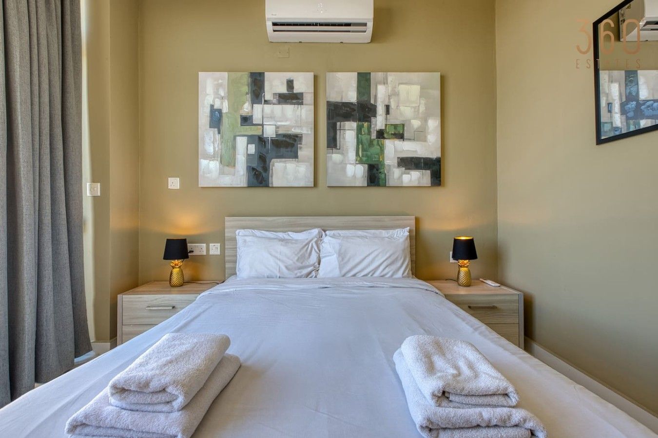 Camera d'albergo con un letto, due lampade da comodino, un condizionatore d'aria a parete e un quadro astratto.