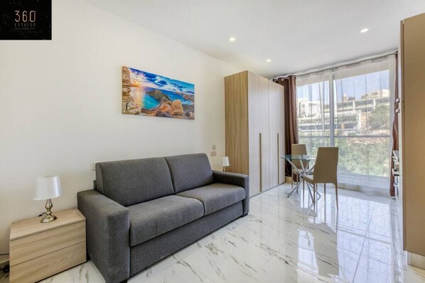 A modern living room with a grey sofa, a glass table, and a painting of a coastal scene.
