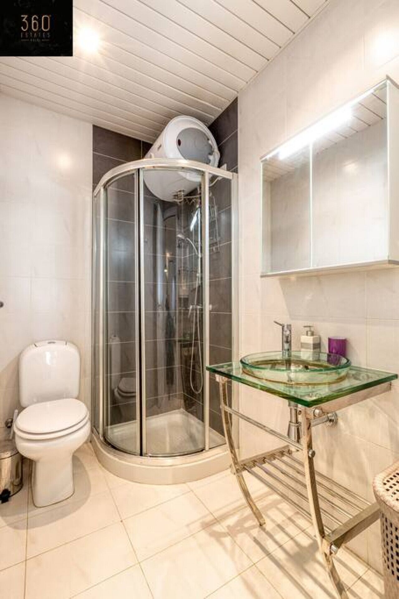 Un baño con ducha de vidrio, inodoro blanco y lavamanos de vidrio con estructura metálica.