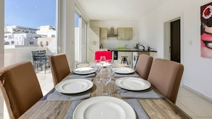 Comedor con una mesa lista para cuatro, cocina al fondo y vista a edificios a través de amplios ventanales.