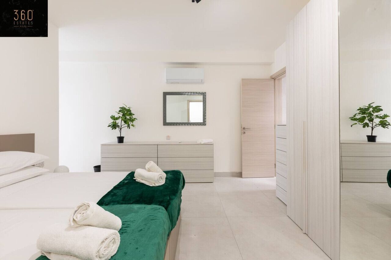 Una habitación de hotel moderna con una cama verde, ropa de cama blanca, un tocador de madera, un espejo y dos plantas en macetas.