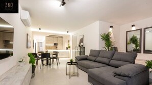A modern living room with a grey sofa, a dining area with a table and chairs, and a kitchen in the background.