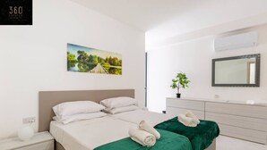 A modern hotel room with a bed, a green throw blanket, a potted plant, a mirror, and a painting on the wall.