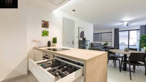 A modern kitchen with a built-in dishwasher, a dining area with a round table and chairs, and a living area with a sofa and a TV.