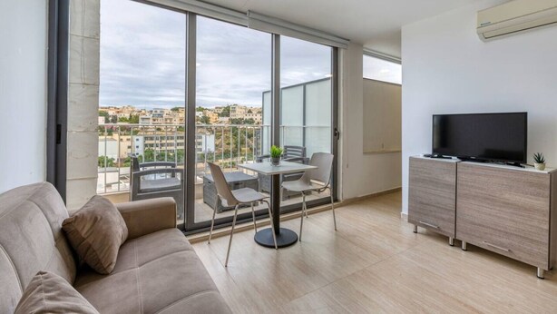 A modern living room with a sofa, a dining table with chairs, a TV on a cabinet, and a balcony with a view of the city.