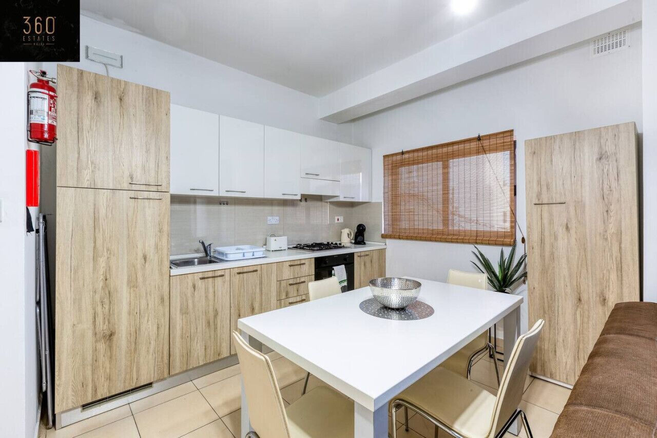 A modern kitchen with white cabinets, a wooden accent wall, a dining table with chairs, and a window with blinds.