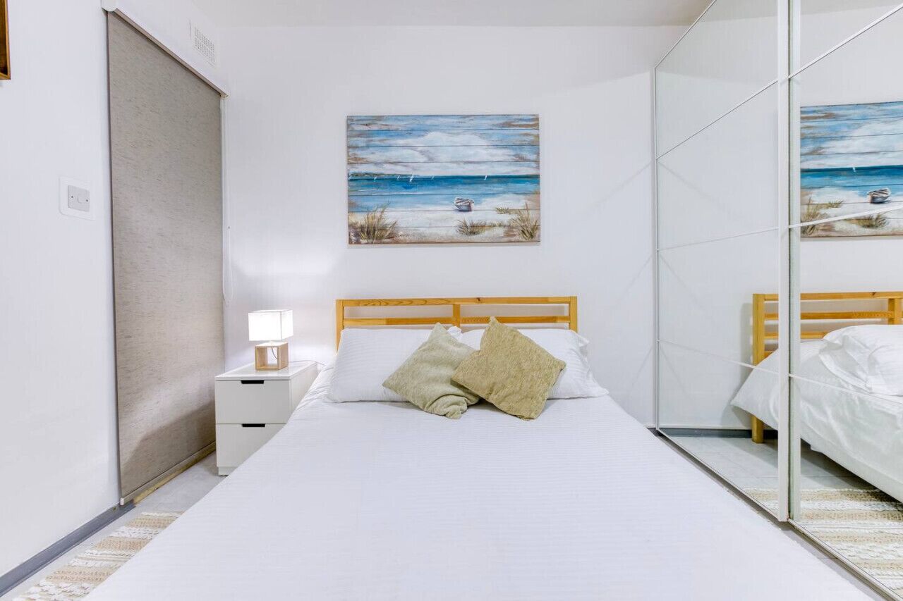 Een slaapkamer met een houten hoofdbord, een witte spreideken, twee kussens, een nachtkastje met een lamp en een schilderij van een zandstrand aan de muur.