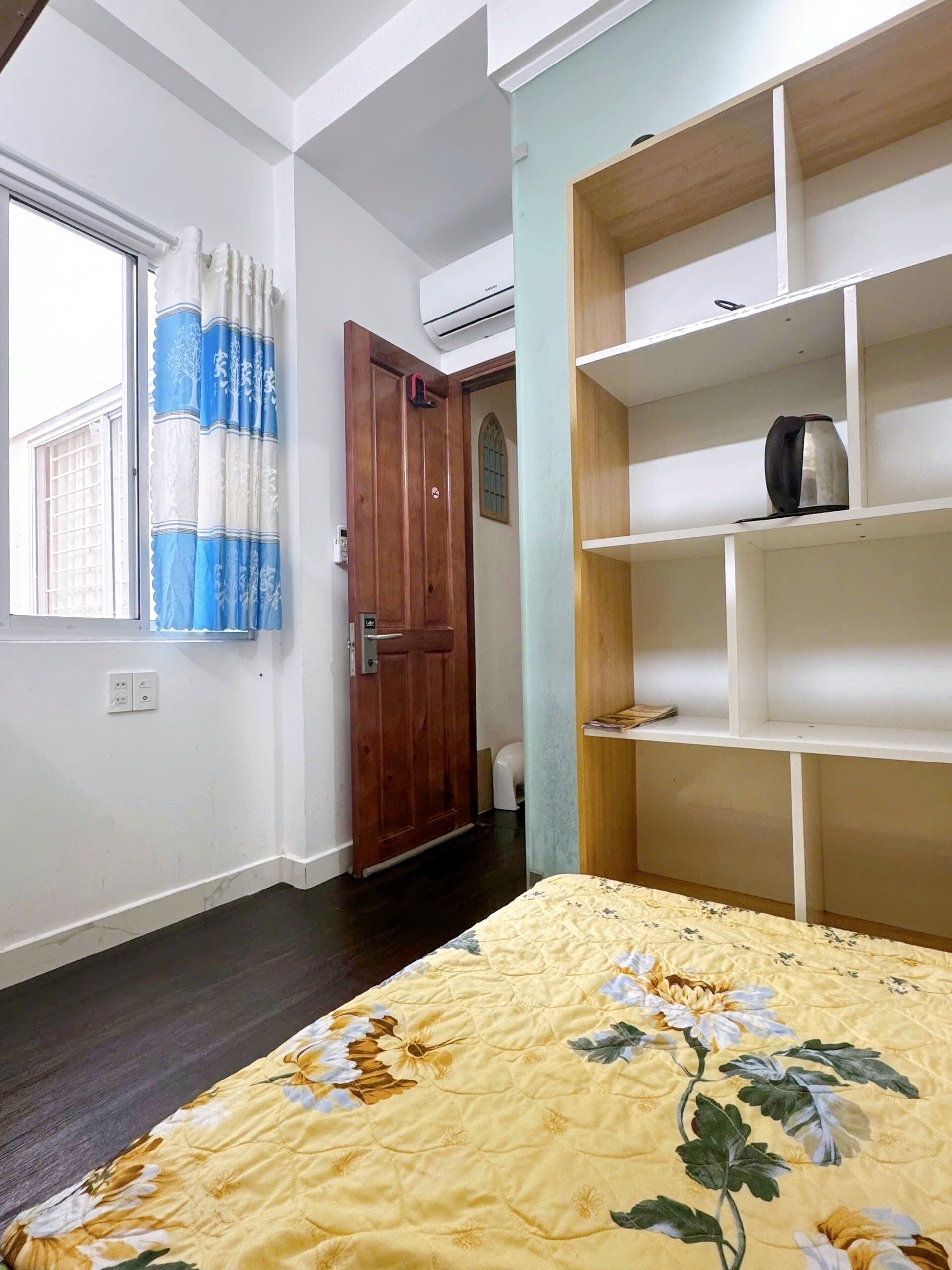 A bedroom with a bed, a window with curtains, a wooden door, and a white shelf unit.