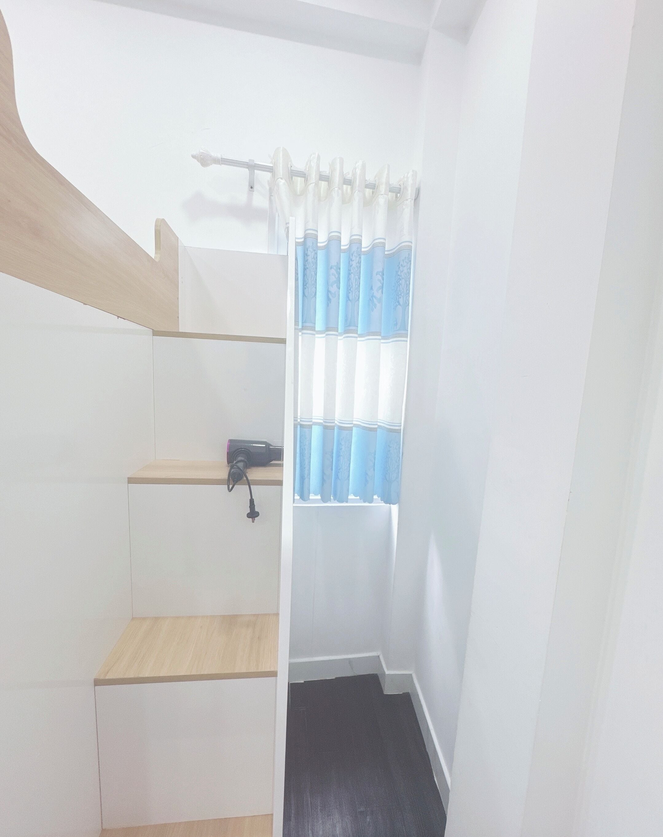 Una escalera con barandilla de madera, un pequeño estante con un objeto negro y una ventana con cortinas azules y blancas.