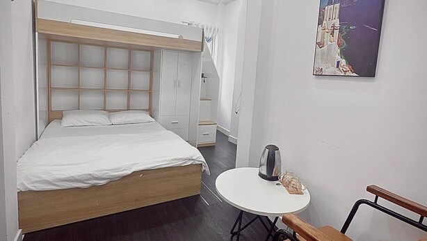 Una habitación de hotel moderna con una cama grande, una mesita redonda con un hervidor, una silla y un cuadro de una escena costera.