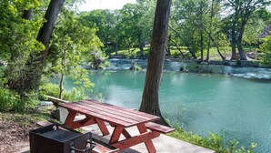 A picnic table with a grill, overlooking a calm river surrounded by trees.