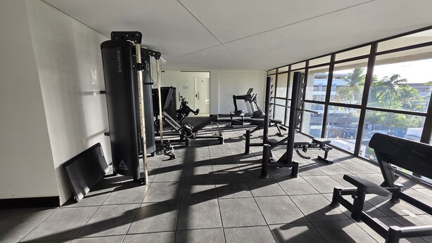 Une salle de sport équipée de divers appareils d’exercice, dotée de grandes fenêtres et d’un sol carrelé.