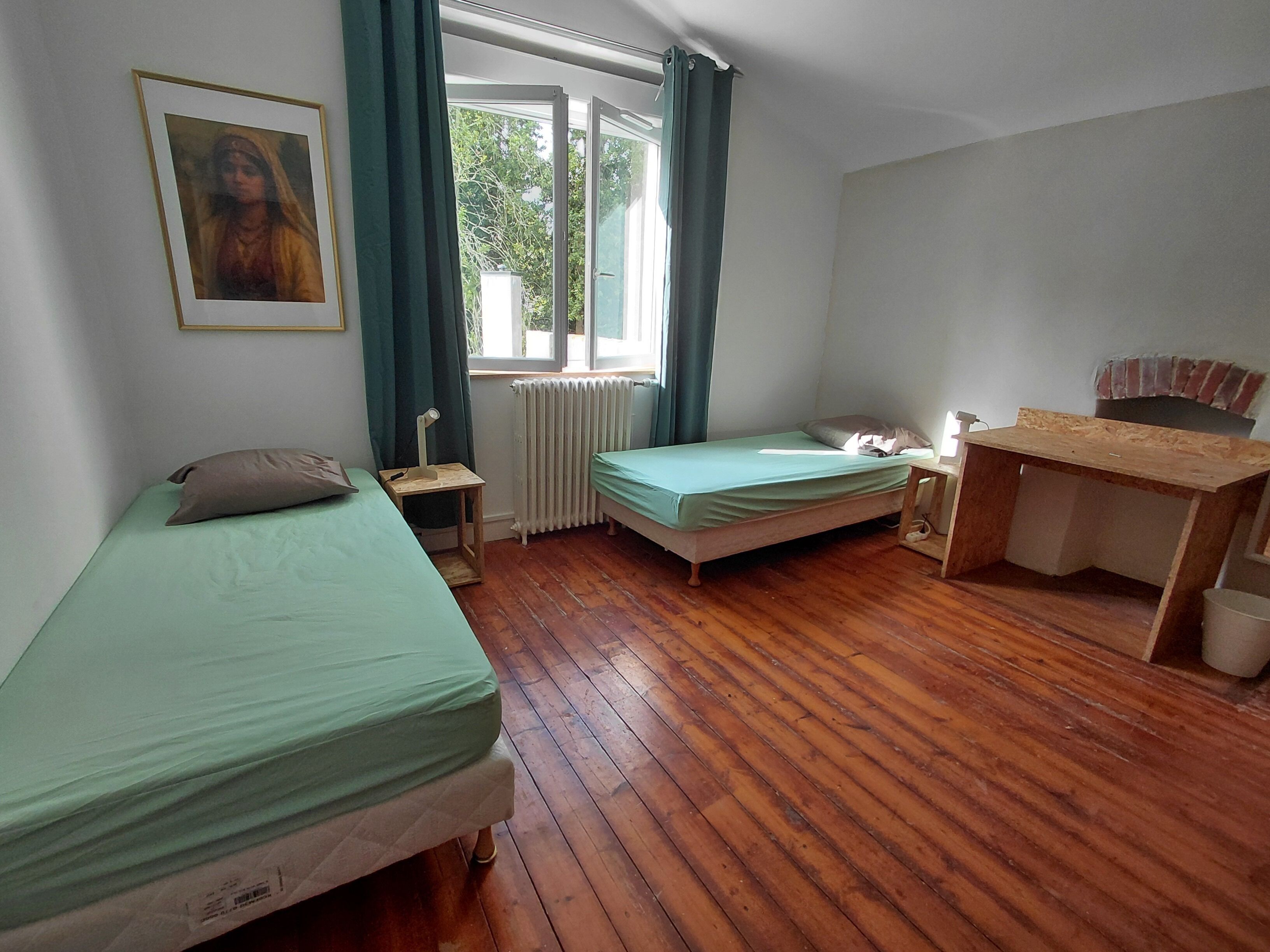 A room with two single beds, a wooden desk, a radiator, and a window with curtains.