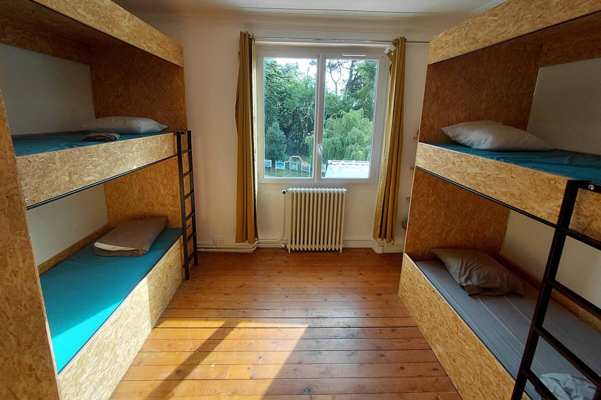Een studentenkamer met stapelbedden, een raam met gordijnen, een radiator en uitzicht op bomen.