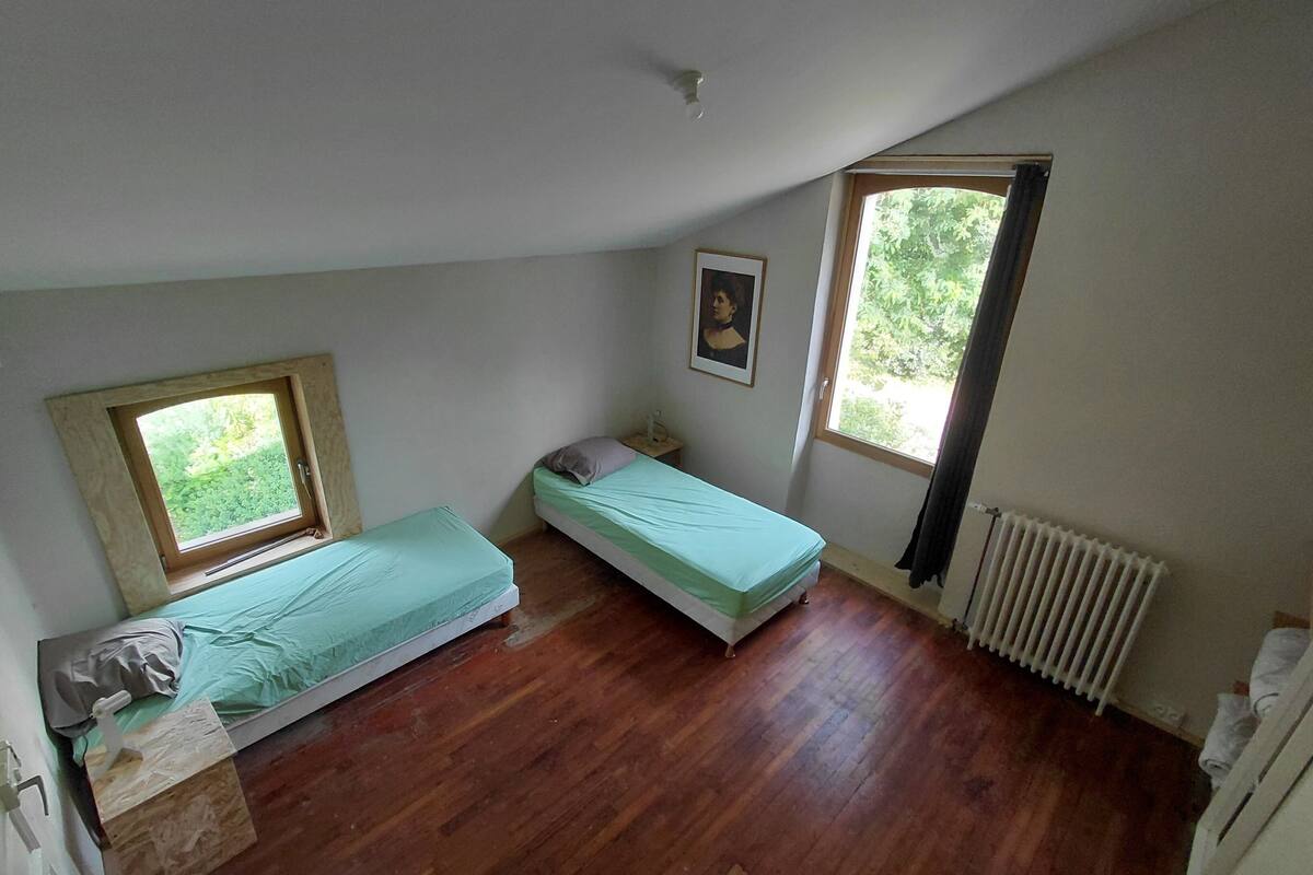 Een klein appartement met één slaapkamer, twee bedden, een radiator en een raam met uitzicht op groen.
